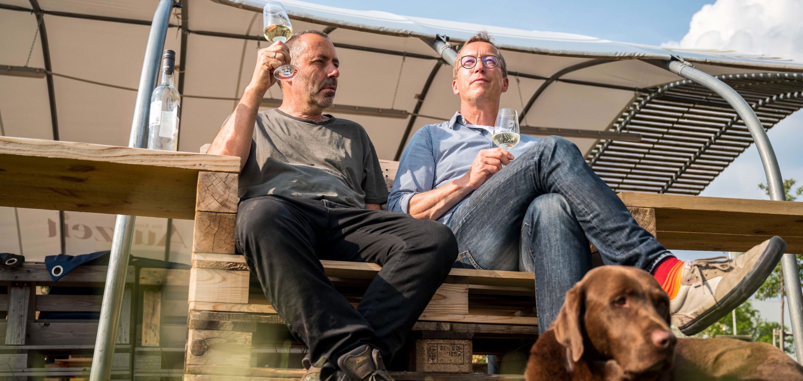Dirk Klöppinger und Georg Merlau sitzen auf Paletten mit Weißwein in der Hand